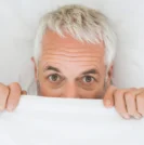 Mature man in bed partially covering face with sheets